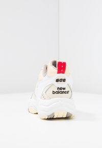 Baskets blanches et beiges avec un dessus texturé, dotées d'une languette rouge au talon, d'une marque noire et d'une semelle en caoutchouc épaisse et jaunie.