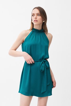 Kvinde iført teal ærmeløs halter-neck kjole med flæset krave og taljebånd, der kigger til siden mod en ensfarvet baggrund.