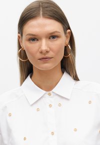 Camicia bianca con colletto, dotata di bottoni decorativi dorati e dettagli sulla parte anteriore, texture del tessuto liscia e design classico.
