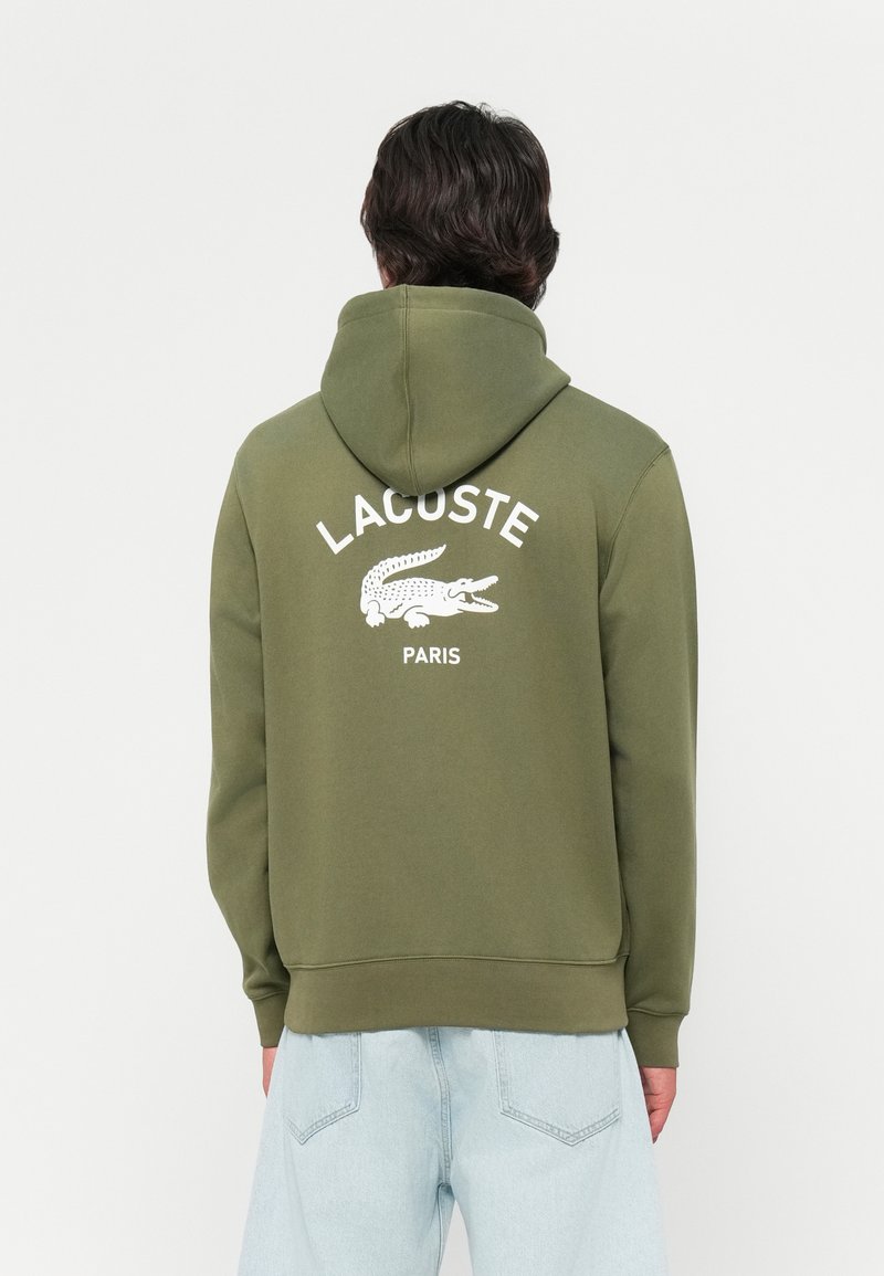 Olijfgroene hoodie met witte "LACOSTE"-tekst en krokodillenlogo op de achterkant, gemaakt van zachte, gestructureerde stof.