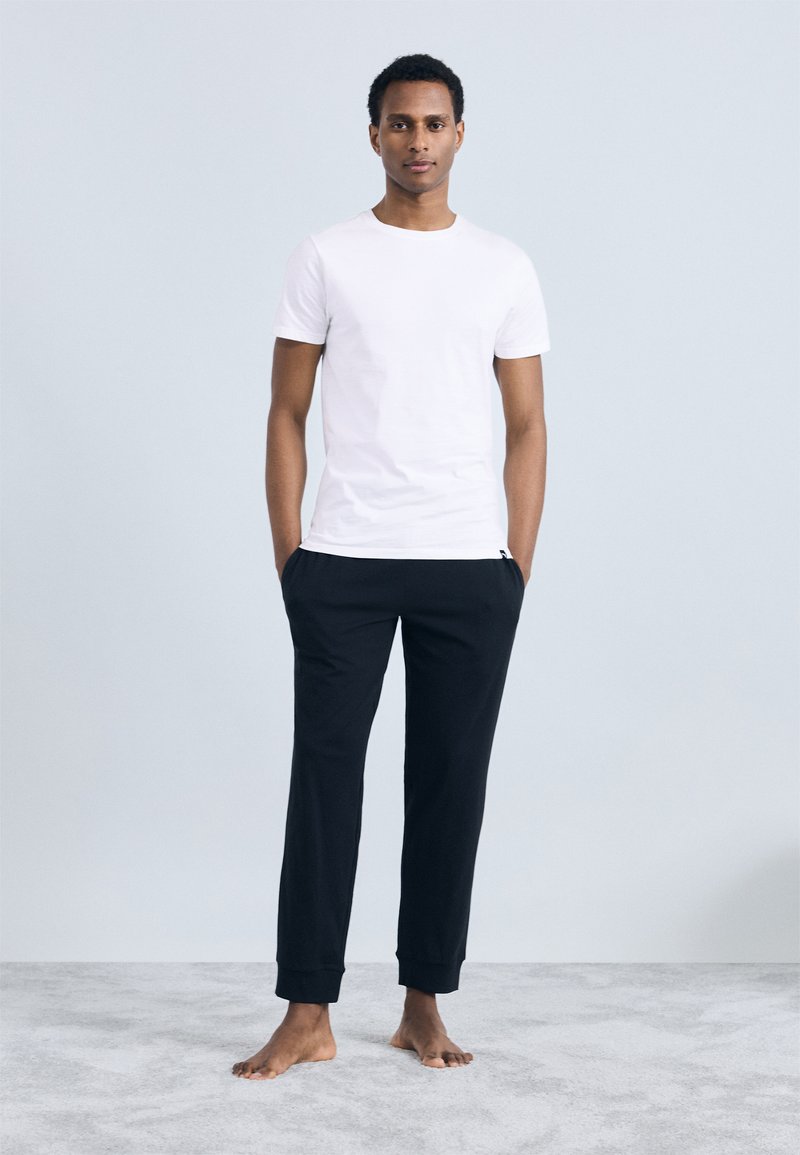 Férfi mezítláb áll a világos szőnyegen, sima fehér pólót és fekete melegítőnadrágot visel, keze a zsebében egy semleges háttér előtt.