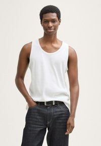 Tânăr îmbrăcat cu un tricou alb fără mâneci și pantaloni negri cu o curea neagră, stând cu o mână în buzunar și un fundal neutru.