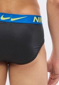 Κοντινό πλάνο ενός ατόμου που φοράει μαύρα εσώρουχα Nike με μπλε ζώνη μέσης που φέρει κίτρινο λογότυπο Nike και κείμενο.