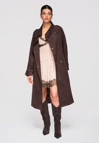 Femme portant un long manteau marron foncé sur une robe beige avec bordure en dentelle noire et des bottes assorties marron foncé montant jusqu'aux genoux, debout devant un fond uni.