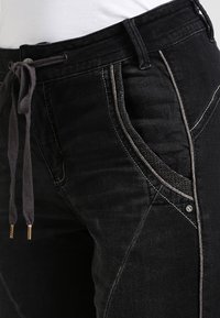 Pantalones de mezclilla negros con cintura ajustable mediante cordón, que presentan costuras en contraste y bolsillos laterales en ángulo con acentos decorativos.