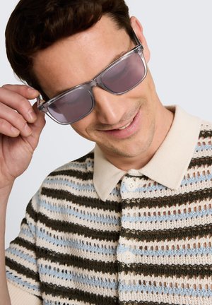 Jonge man glimlacht terwijl hij zijn zonnebril met doorzichtig montuur verstelt, gekleed in een gestreepte gebreide polo in beige, bruin en lichtblauw.