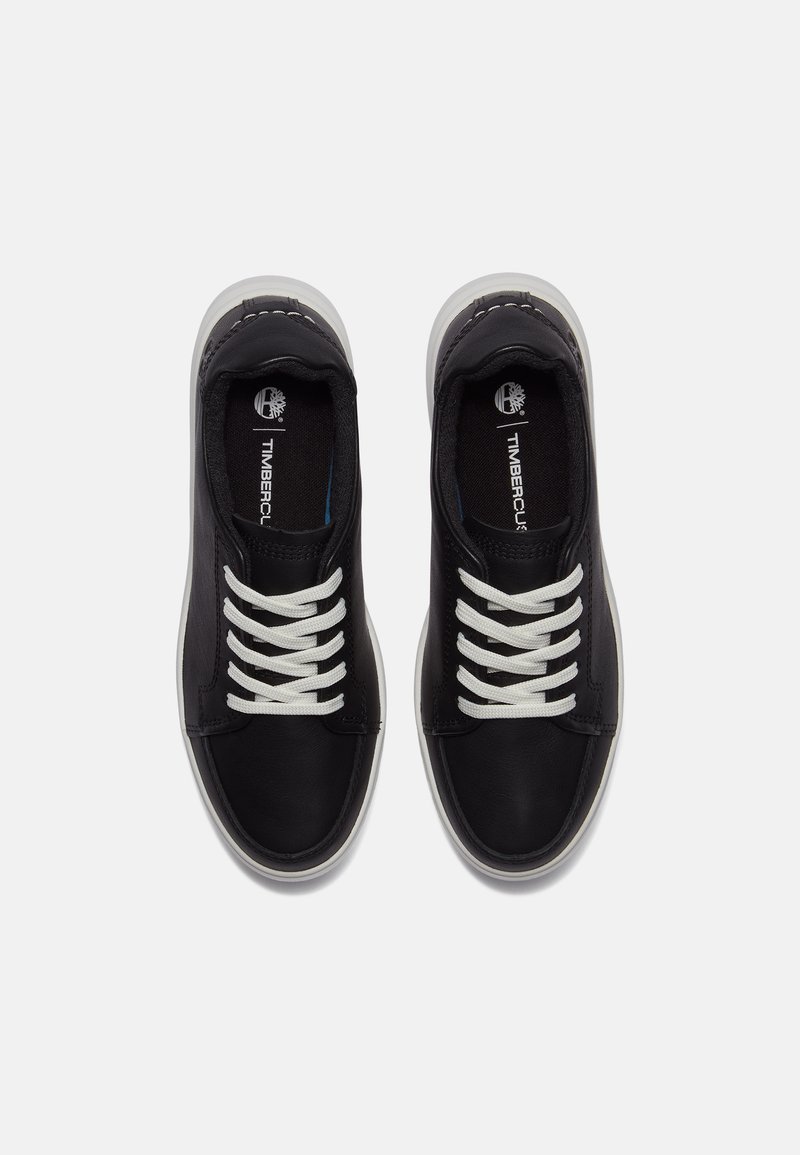 Zapatillas de cuero negro con cordones blancos, punta redondeada y suela de goma gris. Logo de Timberland visible en la plantilla. Textura suave.