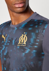 Camisa deportiva Puma en gris oscuro con un patrón texturizado azul, con el logo dorado de Marsella y el texto "DROIT AU BUT" en el frontal.