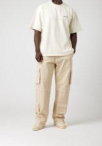 Krämfärgad T-shirt med "EIGHTYFIVE" tryckt på vänster bröst, tillsammans med beiga cargobyxor med sidofickor och lös passform.