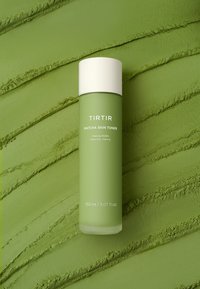 Grüne zylindrische Flasche des TIRTIR Matcha Skin Toners mit weißer Kappe auf strukturiertem grünem Hintergrund, 150 ml, ausgleichende und beruhigende Formel.