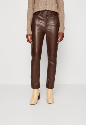 Personne portant un pantalon en cuir brun foncé ajusté, un pull en maille beige et des bottines à talons crème, debout devant un fond blanc uni.