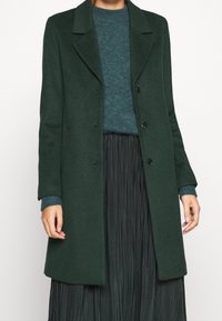 Vrouw in een donkergroene knopenjas over een teal trui en een zwarte geplisseerde rok, staand tegen een effen lichte achtergrond.