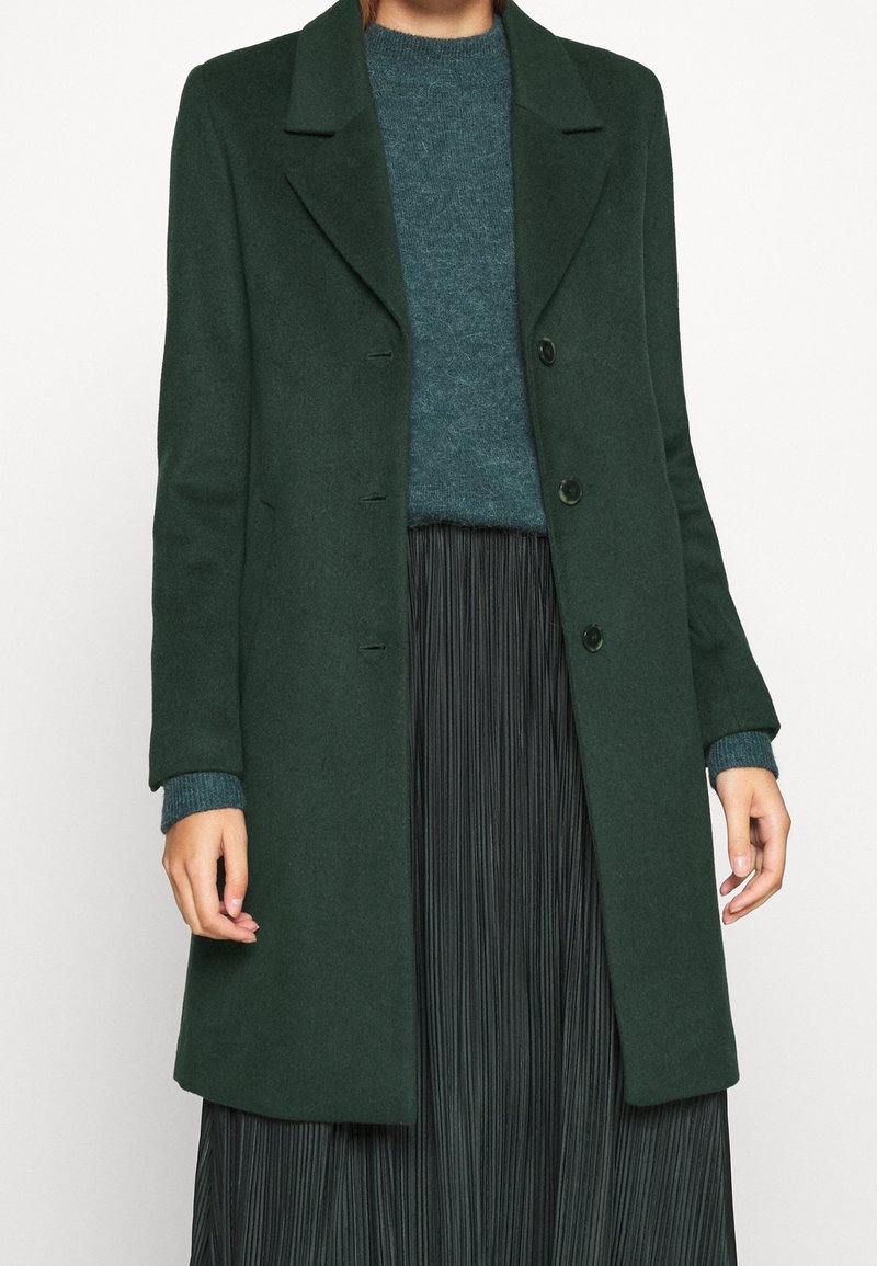 Vrouw in een donkergroene knopenjas over een teal trui en een zwarte geplisseerde rok, staand tegen een effen lichte achtergrond.