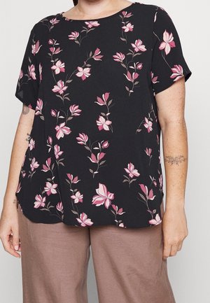 Zwarte blouse met korte mouwen en roze bloemenprint, gedragen met lichtbruine broek, met blote onderarmen met tatoeages en sproetjes.