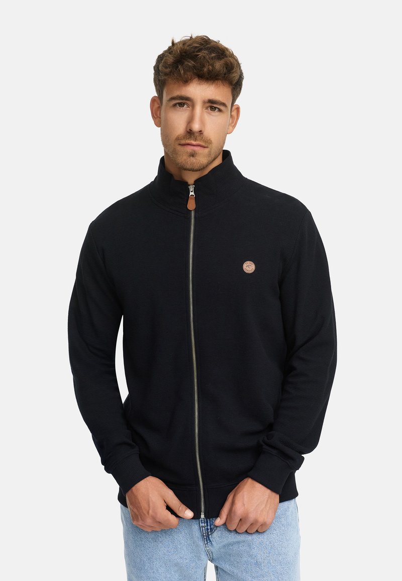 Hombre con cabello rizado corto que lleva una chaqueta negra con cremallera y un pequeño logo redondo en el lado izquierdo del pecho, y jeans de color azul claro.