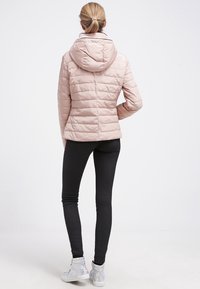 Kvinna med blont hästsvans som bär en ljusrosa vadderad jacka med huva, svarta leggings och silverfärgade högstövlar, stående med ryggen mot kameran.