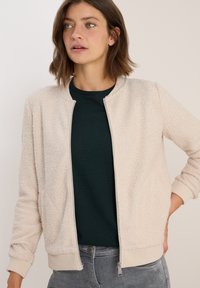 Frau in einer cremefarbenen, strukturierten Zip-Jacke über einem dunkelgrünen Shirt, die mit einer Hand in der Hüfte vor einem schlichten Hintergrund steht.
