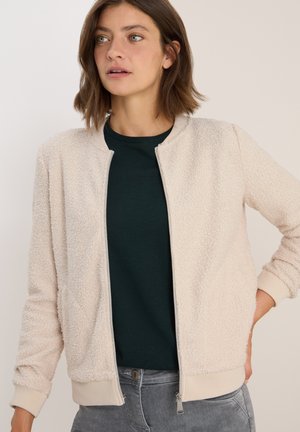 Woman wearing a cream textured zip-up jacket over a dark green shirt, standing with one hand on hip against a plain background.