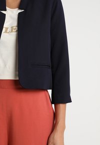 Femme portant un blazer court bleu marine sur un haut blanc avec un texte doré, associé à un pantalon taille haute de couleur rouille.
