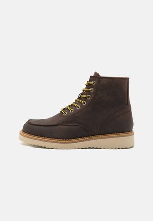 Botas de tobillo de cuero marrón con cordones amarillos, suelas de goma beige, acentos cosidos y diseño de punta redondeada. Textura suave en todo.