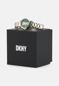 Ceas din metal auriu și argintiu cu un cadran rotund verde, având logo-ul DKNY, expus pe o cutie neagră cu branding alb.