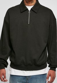 Svart tröja med krage, halvmatchande dragkedja framtill, sänkta axlar och ribbstickade ärmslut. Lagdas över en vit t-shirt.