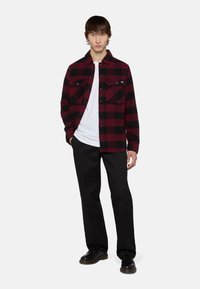 Camisa de franela a cuadros rojos y negros con dos bolsillos en el pecho, botones negros, sobre una camiseta blanca, combinada con pantalones y zapatos negros.