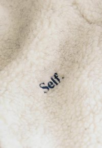 Cremfarbener Fleece-Stoff mit einer weichen, strukturierten Oberfläche, die dunkle blaue, gestickte Schriftzüge mit der Aufschrift "Self" zeigt. Eingebettete Fasern sind sichtbar.