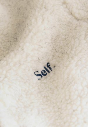 Cream-colored fleece fabric with a soft, textured surface featuring dark blue embroidered text reading "Self." Embedded fibers visible.