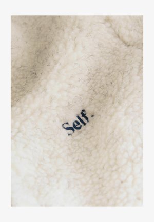 Cream-colored fleece fabric with a soft, textured surface featuring dark blue embroidered text reading "Self." Embedded fibers visible.
