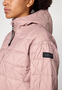 Veste rembourrée matelassée rose clair avec capuche, coutures diagonales et un écusson de marque noir sur la manche. Texture lisse et brillante.