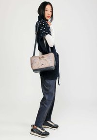 Bolso cruzado en beige con detalles en negro, que presenta patrones intrincados y un cierre superior. Fabricado en material similar al cuero, con un diseño estructurado.
