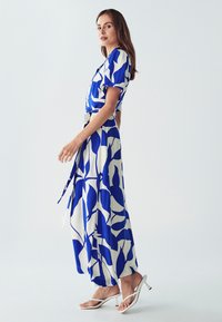 Maxime dress in white with bold blue leaf patterns, featuring short sleeves, a belted waist, and flowing fabric. Paired with white heeled sandals.