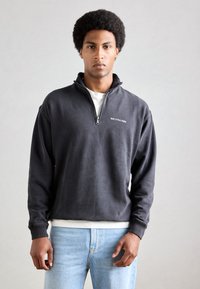 Grå zip-up sweatshirt lavet af blødt materiale, med et lille logo på brystet og ribbede manchetter, parret med lyseblå jeans.