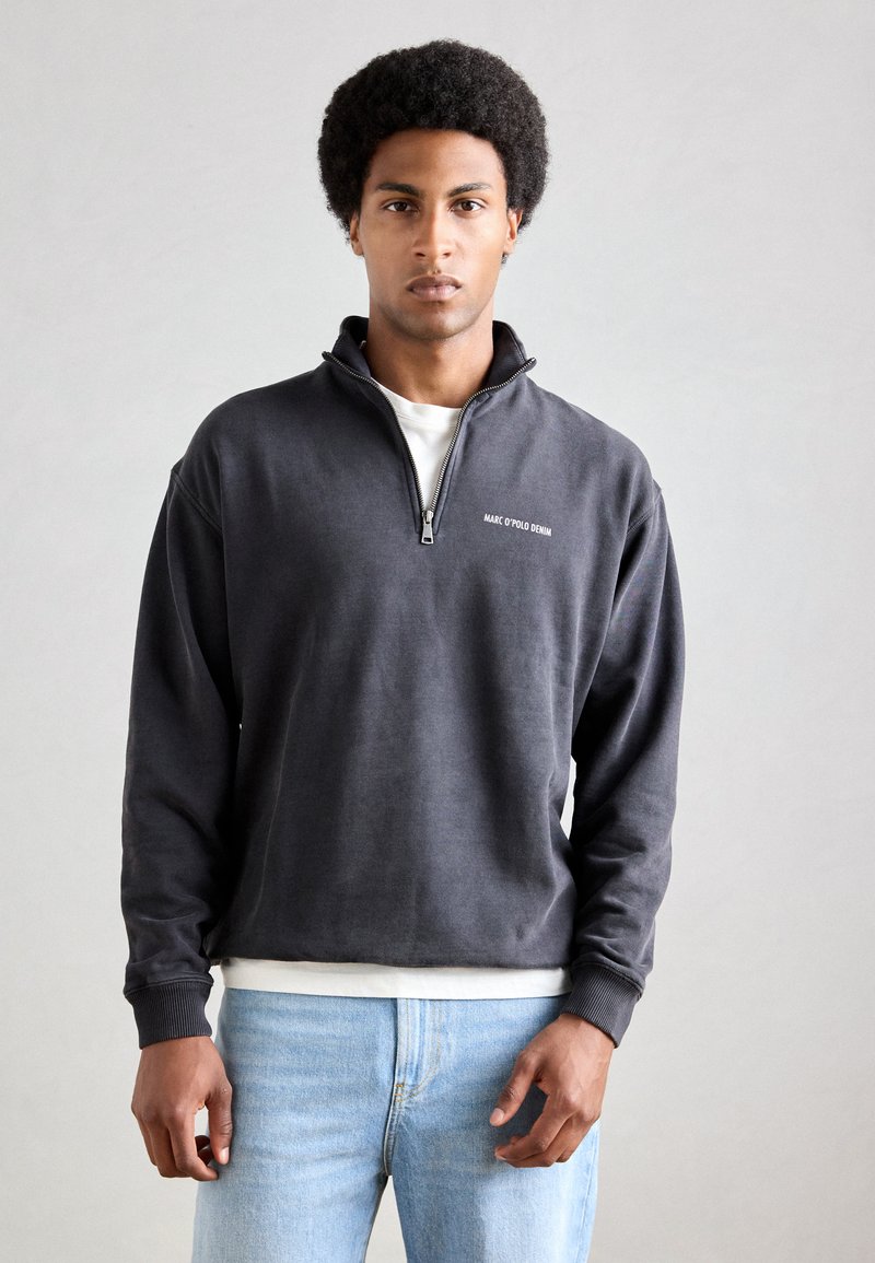 Grå zip-up sweatshirt lavet af blødt materiale, med et lille logo på brystet og ribbede manchetter, parret med lyseblå jeans.