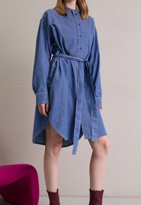 Denim shirt dress with long sleeves, button closure, and a belted waist. Features a rounded hem and subtle contrasting stitching.