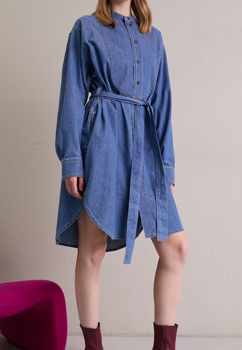 Denim shirt dress with long sleeves, button closure, and a belted waist. Features a rounded hem and subtle contrasting stitching.