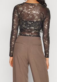 Transparente schwarze Longsleeve-Bluse mit goldenen Sprenkeln, runder Halslinie und taillierter Passform, kombiniert mit hoch taillierten braunen Hosen.