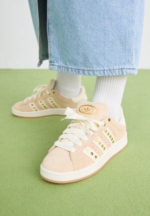 Personne portant des chaussettes blanches, un jean large bleu clair et des baskets Adidas beiges avec des accents dorés, debout sur une surface verte.