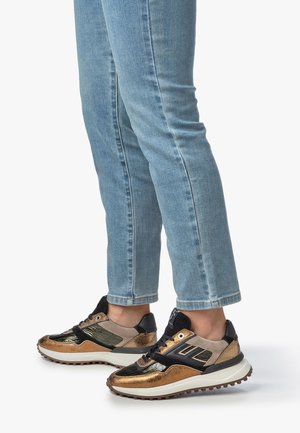 Sneakers featuring a mix of metallic gold, black, and beige materials; textured panels and a chunky sole. Worn with light blue jeans.