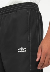 Juodos sportinės šortai „Umbro“ su baltais siūlais, elastinga juosmens juosta, šonine kišenė ir ranka, ilsinanti ant nešėjo klubo.