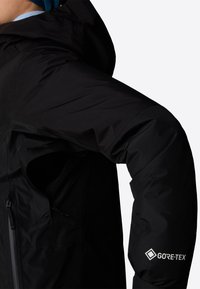 Czarna kurtka GORE-TEX ma teksturowane wykończenie, regulowany kaptur i kieszeń na zamek, z akcentem logo na dolnym rękawie.