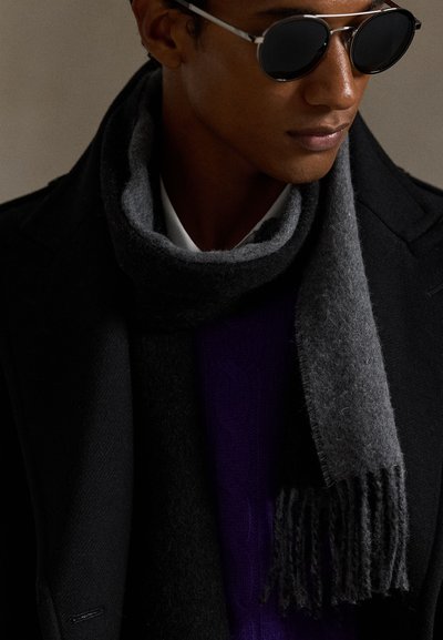 Man wearing dark sunglasses, a black coat, and a thick dark gray scarf wrapped around his neck against a plain background.