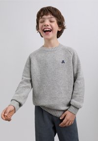 Suéter gris de punto con puños y dobladillo acanalados, con un pequeño emblema azul de calavera en el pecho. Combinado con pantalones azul marino texturizados.