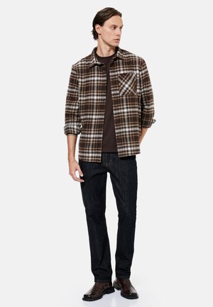 Mand iført en brun ternet skjorte med knapper over en mørk t-shirt, mørke jeans og brune lædersko, stående med den ene hånd bag ryggen.