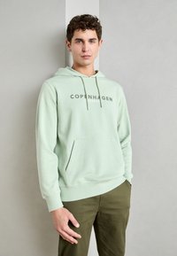 Felpa verde chiaro con tasca a marsupio e cappuccio con cordino. Presenta la scritta "COPENHAGEN LUND" stampata in una tonalità più scura sul petto.
