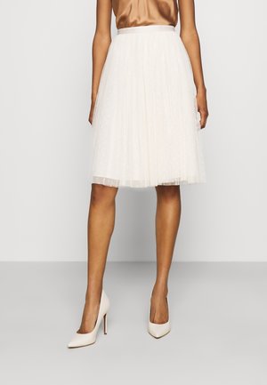 Femme portant une jupe plissée blanche mi-genoux avec une superposition en voile à pois, un haut en satin marron et des chaussures à talons blancs, debout sur un sol gris.