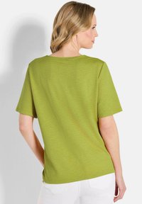 Grünes, strukturiertes Oberteil mit kurzen Ärmeln und Rundhalsausschnitt, kombiniert mit weißen Hosen, von hinten betrachtet.