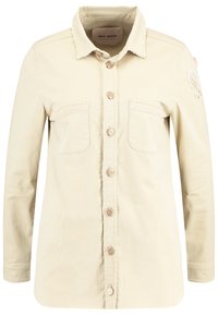 Beige skjorta med knappar i mjukt tyg, med två bröstfickor, broderad bladdetalj på axeln och en fransad krage.