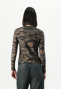 Frau mit glattem schwarzen Haar, die ein langärmliges, enganliegendes Oberteil mit braunem und schwarzem abstraktem Muster und lockeren dunklen Denim-Jeans trägt, Rückansicht.